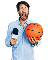 Hispanic man with blue eyes holding reporter microphone and basketball ball angry and mad screaming frustrated and furious, shouting with anger looking up.