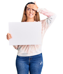 Beautiful young woman holding blank empty banner stressed and frustrated with hand on head, surprised and angry face