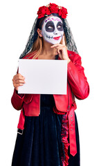 Woman wearing day of the dead costume holding blank empty banner serious face thinking about question with hand on chin, thoughtful about confusing idea