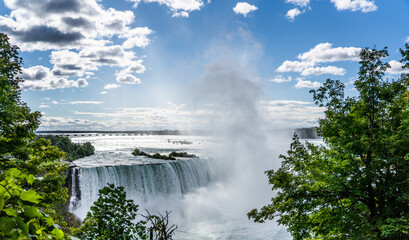 The Horseshoe Falls, part of Niagara Falls on the Canadian site of the Niagara River, partly hidden from green leaves and the sky in partly covered with clouds 