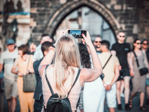 Back view of a blonde unrecognizable woman tourist on Charles Bridge, Prague, taking pictures with her smartphone
