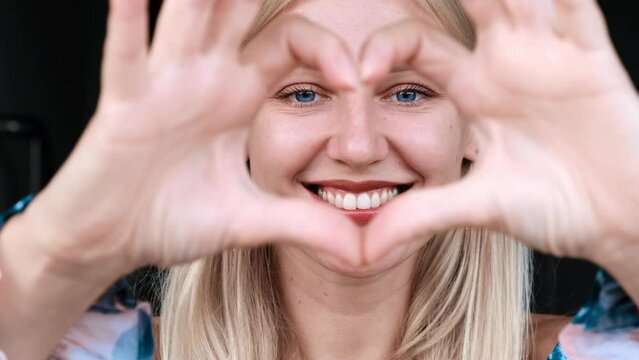 Close-up of happy laughing woman joins her fingers, shows heart symbol on camera, looking at camera, looks overjoyed, says I love you, sincere feelings, sign of affection, provides support and cares