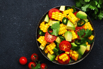 Tex-mex spicy salad with corn, avocado, jalapeno peppers, cherry tomatoes and cilantro. Black stone kitchen table background, top view, copy space
