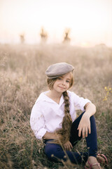 Naklejka na ściany i meble cute little girl with a long braid of blond hair in a white shirt and a men's vintage cap at sunset in a summer field.