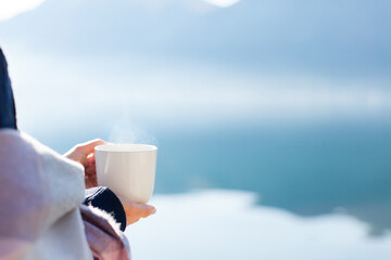 Woman drinking coffee, relaxing at sea beach. Winter picnic with steamy reusable cup by mountains, lake. Girl enjoying ecological travel, slow life, morning tea, peace, calmness. Blue still water