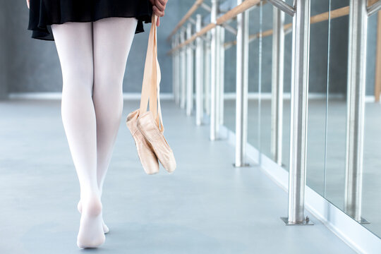 Ballerina taking ballet pointe shoes in hands. Girl finishing barre workout and going barefoot in class room of dance. Close up of legs in studio. Woman has rest and break - Powered by Adobe