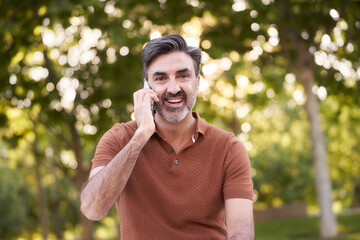 Middle-aged man looking at the camera smiling while talking on the phone outdoors in a park. Technology concept.