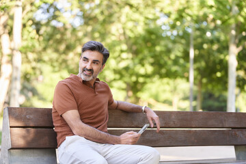 Relaxed man using a mobile phone while sitting outdoors in a park bench. Technology concept.