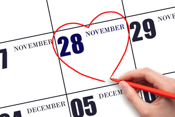 A woman's hand drawing a red heart shape on the calendar date of 28 November. Heart as a symbol of...