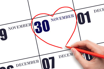 A woman's hand drawing a red heart shape on the calendar date of 30 November. Heart as a symbol of love.