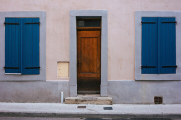 House facade with a dark brown wooden door and two closed blue wooden windows. Frontal view with sidewalk. No people. Traditional house in an old town of France