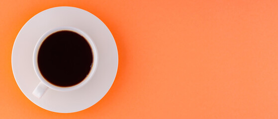 White cup with coffee on saucer on orange coral background. Black coffee for breakfast. Top view. Panoramic shot