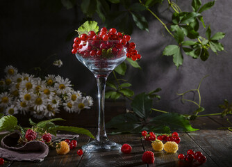 Red currants in a glass wine glass, raspberries and a bouquet of daisies on a wooden table