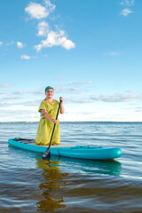 A woman in a dress and a headdress standing on a SUP board with an oar floats on the water against the blue sky.