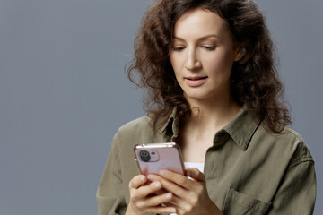 Focused smiling pensive curly beautiful woman in casual khaki green shirt typing new post in blog using phone posing isolated on over gray blue background. Social Media Influencer concept. Copy space
