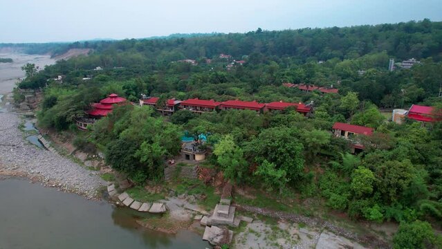 decending aerial drone shot showing the red roofs of resort nestled between green tress with swimming pool and cottages to the kosi dried riverbed located at jim corbett park in India