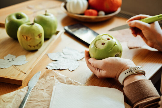 Halloween DIY Fruit ideas. Female hands cutting out Halloween green apple with creepy carved face on wooden table at home. Healthy Halloween Snacks