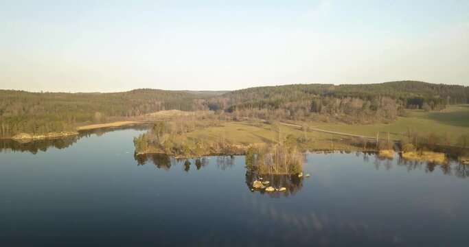 Drone flying over lake surounded bny forest and farmland in early morning 
