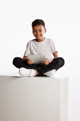 Curly school age boy in casual tee and jeans sitting on big box with digital tablet isolated on...