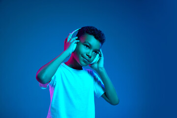Little cheerful happy boy 6-7 years old wearing white t-shirt and headphones listen to music isolated on blue background in neon light.