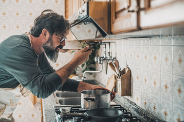 Man at home tasting food from the pot with a table spoon. People in the kitchen preparing dinner. One mature male alone cooking. Real lifestyle person testing taste and cook preparation. Husband work
