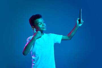 Cute little boy, kid using smartphone isolated over blue background in neon light. Education, childhood, modern lifestyle