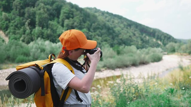 A young woman takes pictures on a DSLR camera. Hiking with camera, orange backpack and cap. A tourist girl takes pictures of nature during a trip. A trip to rest in nature in the forest and mountains.