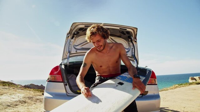 Sporty shirtless man waxing surfboard at the car trunk on sand beach