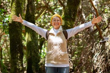 Happy caucasian senior woman hiking in the forest holding backpack enjoying freedom and nature -...
