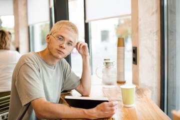 Stylish blond teenage boy 18-19 year old wear glasses reading paper book in cafe with coffee in cup. Young teen student studying.