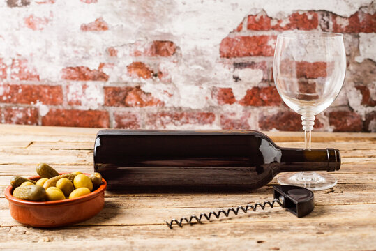 Bottle of red wine lying on an aged wooden table next to a glass, a corkscrew and a plate with olives and pickles, with an old wall background.