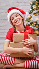 Happy Christmas. Portrait of a Christmas happy smiling girl in a red hat, holding a Christmas gift in her hands and looking into the camera. Sitting in a room on the floor against the background of a