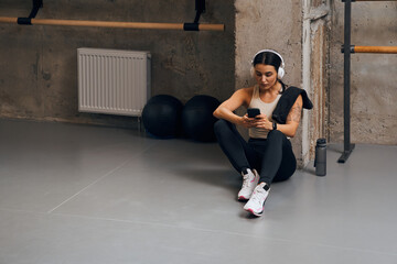 Fototapeta na wymiar Fitness woman sitting in gym while using phone and listening music with earphones