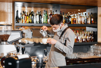A masked barista prepares an exquisite delicious coffee at the bar in a coffee shop. The work of restaurants and cafes during the pandemic.