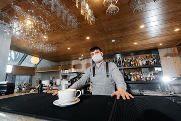 A masked barista exquisitely serves ready-made coffee in a modern cafe during a pandemic. Serving ready-made coffee to a client in a cafe.