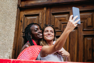 two women friends of different ethnicity take a selfie in the street during the popular festivities...