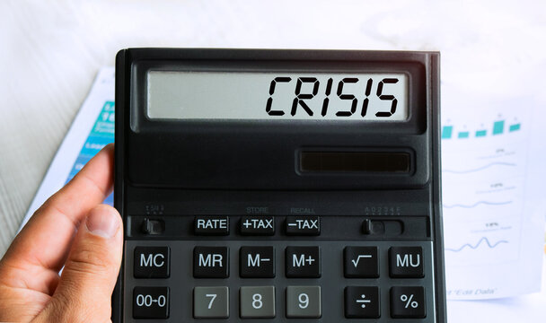 The word CRISIS is written on the calculator. Business man holding a calculator in his hand. Top view of the desktop and business charts. Finance or business concept.