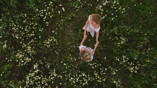 Two young women in summer dresses holding hands and walking in a circle in the field of daises
