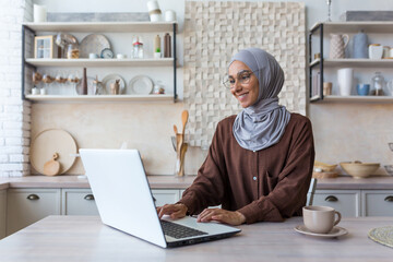 Young beautiful Muslim woman in hijab working remotely from home, using laptop while sitting in kitchen at table, woman in glasses smiling and happy with work.