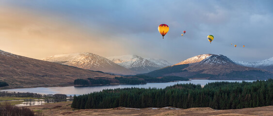 Digital composite image of hot air balloons flying over Majestic beautiful Winter sunrise panorama landscape image of glowing light on mountain range and peaks beyod Loch Tulla in Scottish Highlands