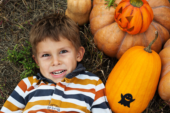 Portrait of cute preschool boy, lies on grass next to pumpkins and makes faces. Nature, outdoor, October day. Happy Halloween.