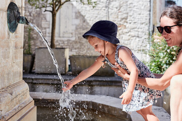 petite fille, tenue par sa m&egrave;re, joue avec l'eau d'une fontaine pour se rafraichir en pleine canicule