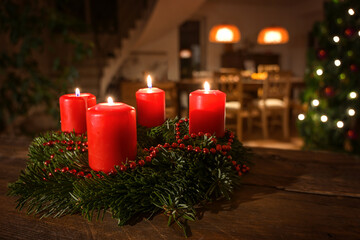 Decorated Advent wreath from fir branches with red burning candles on a dark wooden table, living room with Christmas tree blurred in the background, copy space