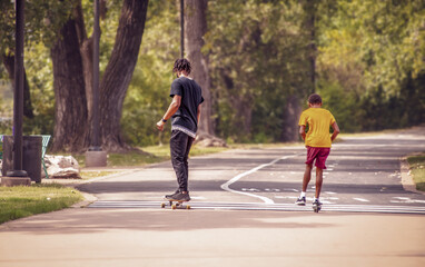 Two black boys one on scooter and one on skateboard on park trail with tall trees on one side -...