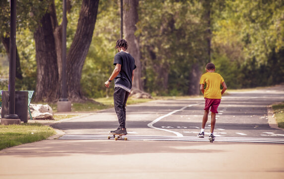 Two black boys one on scooter and one on skateboard on park trail with tall trees on one side - motion blur and facing away from camera