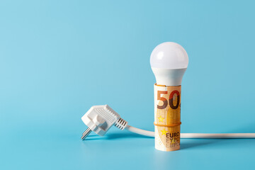 LED light bulb on a rolled 50 euro banknotes and white power plug over blue background. Rising...