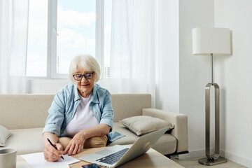 an elderly blonde woman in stylish clothes is sitting on the couch at home and working on a laptop...