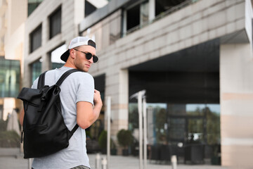 Handsome young man with stylish sunglasses and backpack near building outdoors, space for text - obrazy, fototapety, plakaty