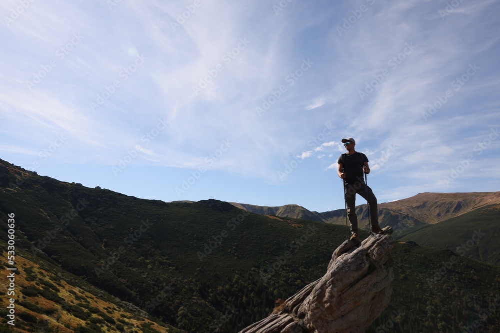 Wall mural man with backpack and trekking poles on rocky peak in mountains