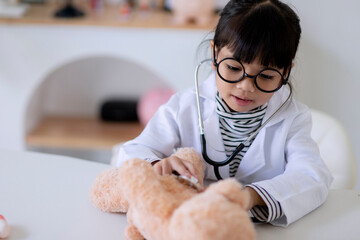 Asian girl playing with her patient plush bear in a doctor game, using a stethoscope. Children want...